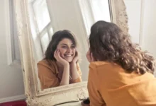 A young woman with curly brown hair and a mustard-colored top smiles warmly at herself in an ornate mirror.