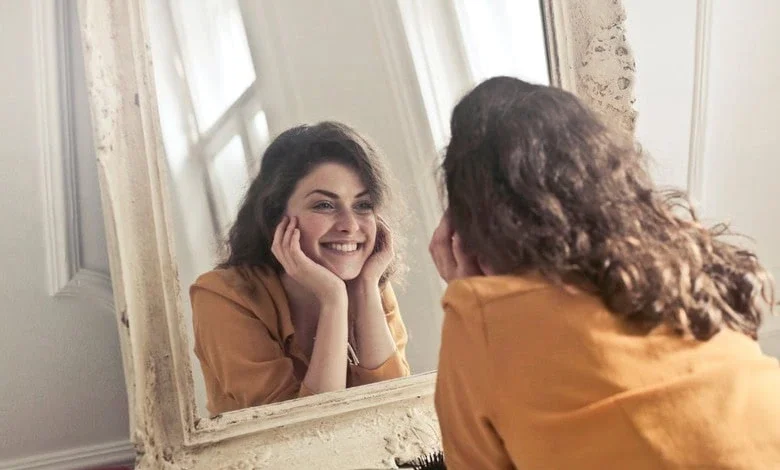 A young woman with curly brown hair and a mustard-colored top smiles warmly at herself in an ornate mirror.