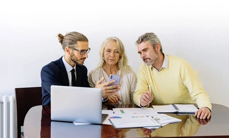 A financial advisor in a suit explains investment details to an elderly couple using a smartphone and documents.