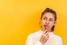 A young woman in a white sweater holds a red toothbrush to her lips, appearing thoughtful against a vibrant yellow background.