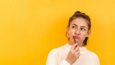 A young woman in a white sweater holds a red toothbrush to her lips, appearing thoughtful against a vibrant yellow background.