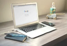 Laptop displaying the Google homepage on a wooden desk with a notebook, pens, smartphone, and a small decorative cannon.