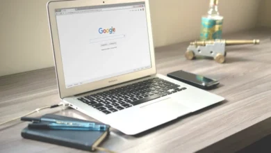 Laptop displaying the Google homepage on a wooden desk with a notebook, pens, smartphone, and a small decorative cannon.