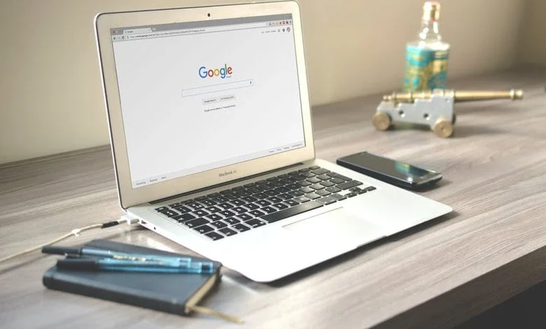 Laptop displaying the Google homepage on a wooden desk with a notebook, pens, smartphone, and a small decorative cannon.